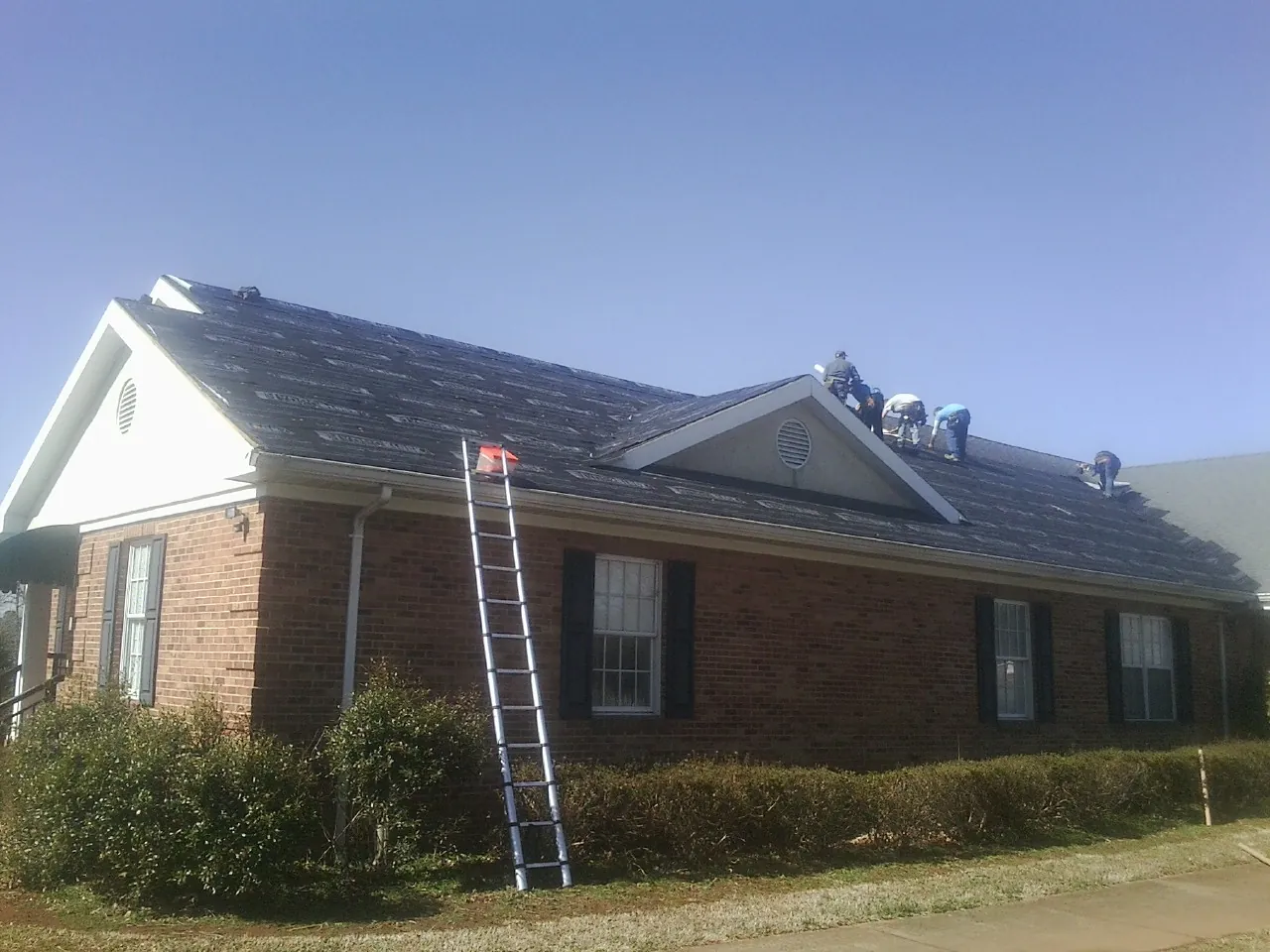 Crew tearing shingles off a brick ranch roof with a ladder set — Metal Roof Repair work by our crew in Hostetter