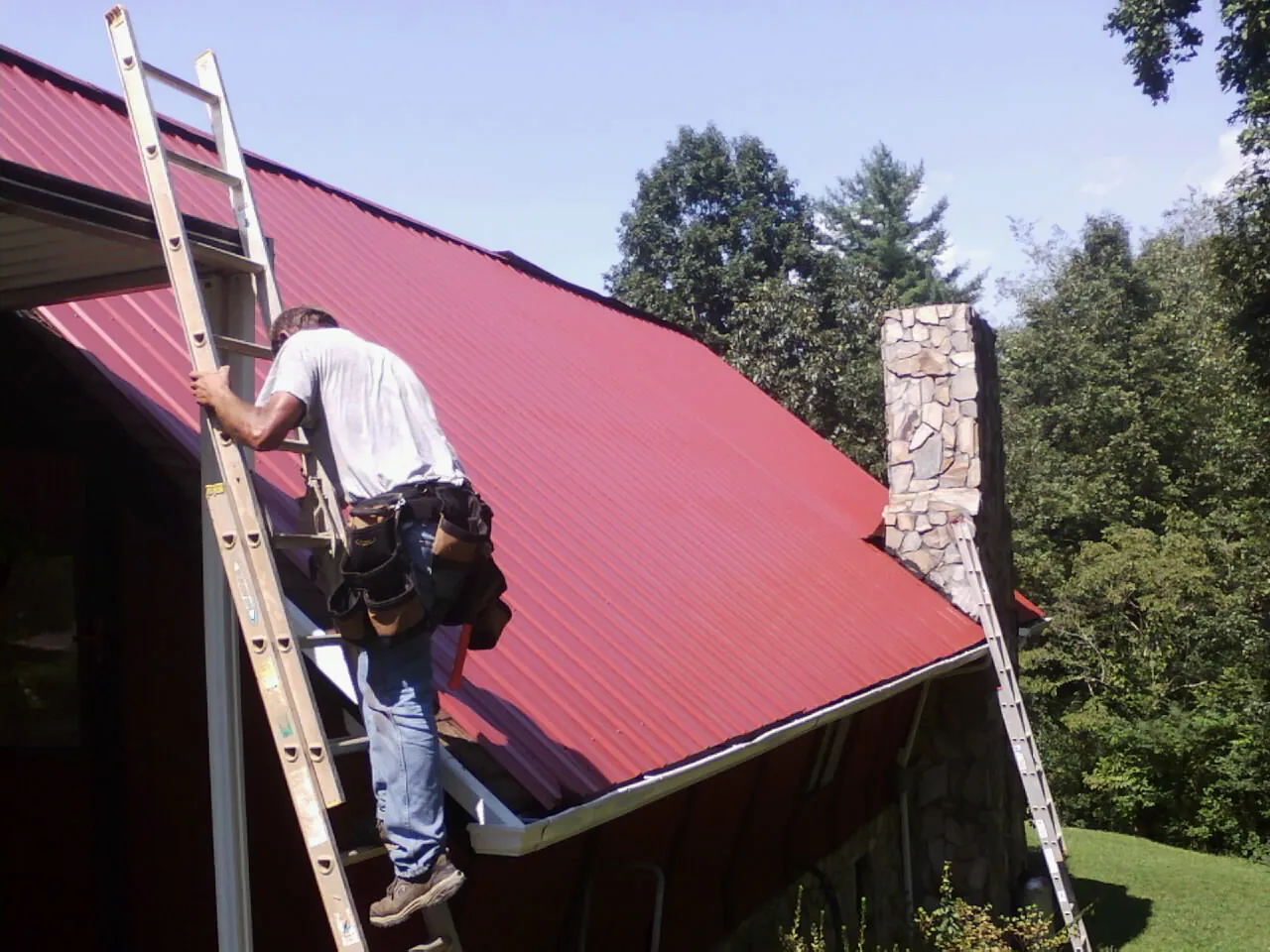 Roofer on a ladder beside a red metal roof and stone chimney — Soffit & Fascia Repair by Roofing Brothers in Hostetter